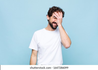 young crazy bearded man laughing and slapping forehead like saying d’oh! I forgot or that was a stupid mistake against flat wall