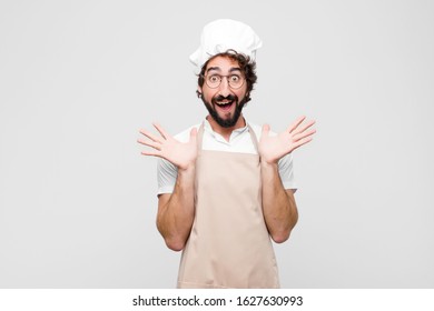 Young Crazy Chef Looking Happy And Excited, Shocked With An Unexpected Surprise With Both Hands Open Next To Face Against White Wall