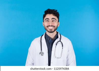 Young crazy doctor smiling with cross eyed wearing the coat hanging the stethoscope around neck on a blue background.