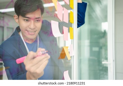 Young Creative Businessman Holding A Marker And Writing Plan And Share Idea On Glass Wall With Sticky Note, Brainstorming And Discussing And Formulating, Business Strategies In Tech Startup Office.