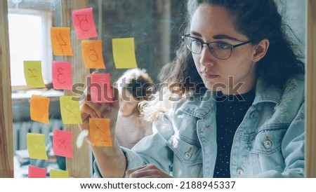 Young creative businesswoman numbering coloured stickers on glassboard for project while working together with female colleague in modern office indoors