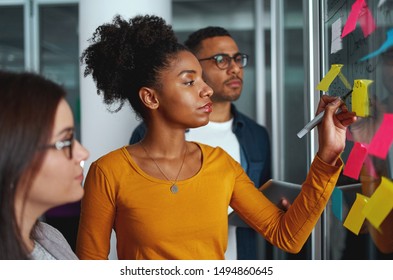 Young creative businesswoman standing with her colleagues writing new ideas on sticky notes over glass wall - Powered by Shutterstock