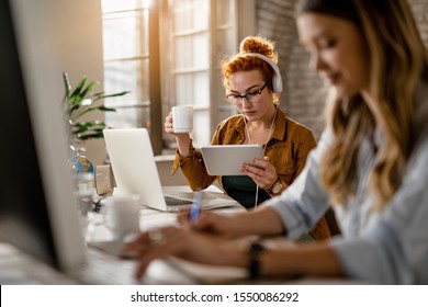 Young creative businesswoman wearing headphones while surfing the net on digital tablet and drinking coffee in the office. 