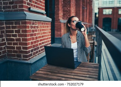 Young creative female freelancer in spectacles enjoying aroma coffee while sitting at wooden table with laptop computer.Talented student drinking tasty beverage during distantly work at street
