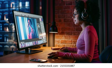 Young Creative Female In Headphones Using Computer In Stylish Loft Apartment In The Evening. Graphic Designer Working From Home, Editing Fashion Photo For Online Store. Urban View From Big Window.