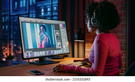 Young Creative Female in Headphones Using Computer in Stylish Loft Apartment in the Evening. Graphic Designer Working from Home, Editing Fashion Photo for Online Store. Urban View from Big Window. - Powered by Shutterstock