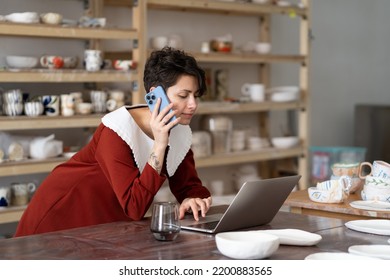 Young creative woman artisan artist ordering materials for ceramic work by phone while working in studio or workshop, female pottery shop owner talking with client via cellphone and typing on laptop - Powered by Shutterstock