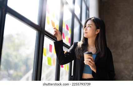 Young creative woman professional using sticky notes in glass wall to writing strategy business plan to development grow to success. Happy asian woman working at office. - Shutterstock ID 2255793739