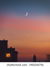 Young Crescent Moon On The Sky Over City. Silhouette Of Towers And Houses With Chimneys On Roofs. Night City Buildings With One Lonely Light In The Window With Flowerpot. Cityscape. Evening Home Scene