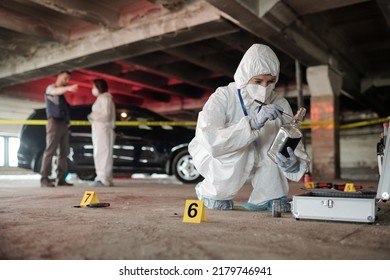 Young criminological expert in coveralls inspecting empty bottle on crime scene while squatting in front of open briefcase with working supplies - Shutterstock ID 2179746941