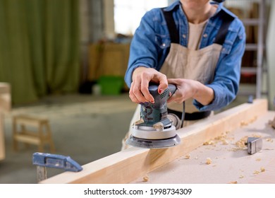 Young Cropped Female Carpenter Working With Wood Using Electric Sander In Workshop. Joinery Work On The Production And Renovation Of Wooden Furniture. Small Business Concept. Close-up Photo