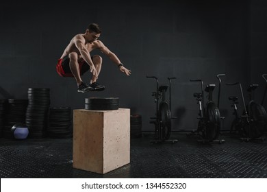 Young Crossfit Athlete Doing Box Jump Exercise At The Gym. Man Jumping Onto The Box On Gray Wall Background. Copy Space