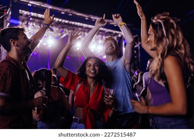 A young crowd of multiethnic friends are dancing at a concert having beers and holding their hands up in their casual clothing.
