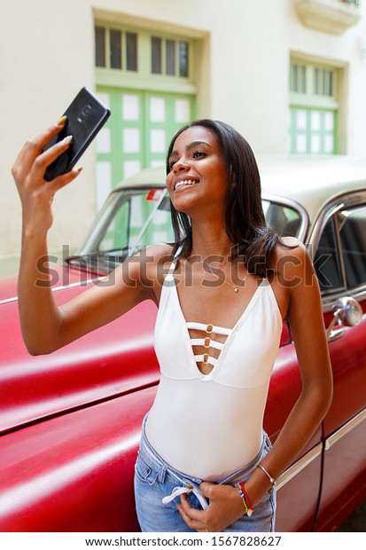 Young Cuban taking a selfie in front of an old car\
in Havana, Cuba