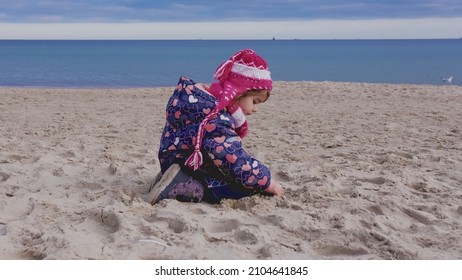 Young Curious Caucasian Girl In Warm Winter Jumpsuit Searching For Seashells And Amber Rock In Beach Sand On Cold Autumn Winter Day