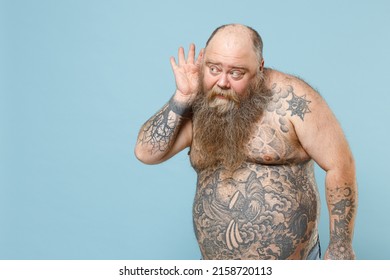 Young curious fun fat pudge obese chubby overweight blue-eyed bearded man 30s has big belly with naked tattooed torso try to hear you overhear listening intently isolated on pastel blue background.