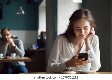 Young Curious Guy Likes Millennial Beautiful Girl Looking With Interest Watching Smiling Lady Sitting Nearby In Cafe Using Smartphone, Flirt In Public Place, Dating And Love At First Sight Concept