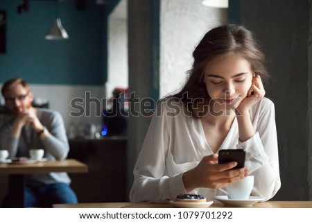Young curious guy likes millennial beautiful girl looking with interest watching smiling lady sitting nearby in cafe using smartphone, flirt in public place, dating and love at first sight concept