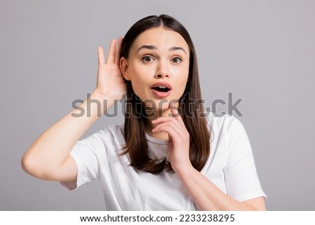 Young curious inquisitive woman girl lady standing on grey background in studio isolated holding hand near ears trying to overhear new information gossip about somebody or discounts.