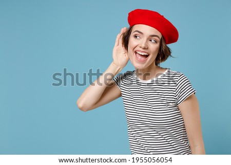 Young curious nosy fun european woman 20s with short hairdo wearing french beret red hat striped t-shirt try to hear you overhear listening intently isolated on pastel blue background studio portrait