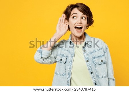 Young curious nosy fun happy woman wearing green t-shirt denim shirt casual clothes try to hear you overhear listening intently isolated on plain yellow background studio portrait. Lifestyle concept