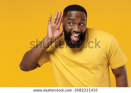 Young curious nosy happy black man 20s wearing bright casual t-shirt try to hear you overhear listening intently isolated on plain yellow color background studio portrait. People lifestyle concept