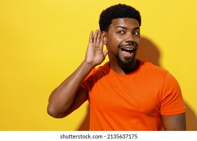 Young Curious Nosy Man Of African American Ethnicity 20s Wear Orange T-shirt Try To Hear You Overhear Listening Intently Isolated On Plain Yellow Background Studio Portrait. People Lifestyle Concept