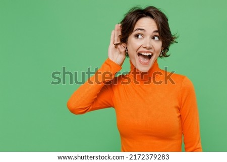 Young curious nosy woman 20s wear casual orange turtleneck try to hear you overhear listening intently isolated on plain pastel light green color background studio portrait. People lifestyle concept