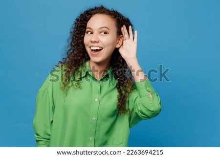 Young curious nosy woman of African American ethnicity 20s she wear green shirt try to hear you overhear listening intently isolated on plain blue background studio portrait. People lifestyle concept