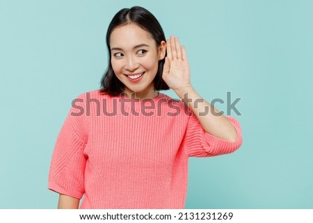 Young curious nosy woman of Asian ethnicity 20s wearing pink sweater try to hear you overhear listening intently isolated on pastel plain light blue color background studio. People lifestyle concept