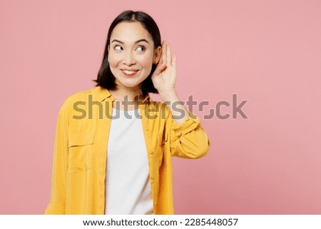 Young curious nosy woman of Asian ethnicity wear yellow shirt white t-shirt try to hear you overhear listen intently isolated on plain pastel light pink background studio portrait. Lifestyle concept