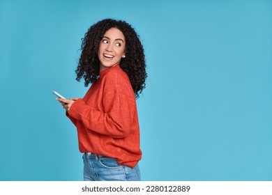 Young curious smiling happy pretty latin woman holding mobile phone, doing online shopping on cell, using apps on cellphone looking aside at copy space standing isolated on blue background. - Shutterstock ID 2280122889