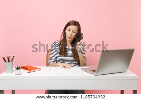 Young Curious woman scrutinizing looking through magnifying glass on pc laptop while sit work on project at office isolated on pastel pink background. Achievement business career concept. Copy space