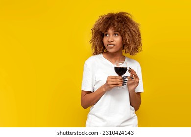 young curly american girl with braces holds glass of wine on yellow isolated background, african woman in white t-shirt with red wine smiles and looks at copy space