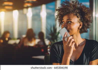 Young curly black female in luxury office space recording audio message via her smartphone, afro american teenage curly girl using her cell telephone as recorder to send voice message to her mother - Powered by Shutterstock