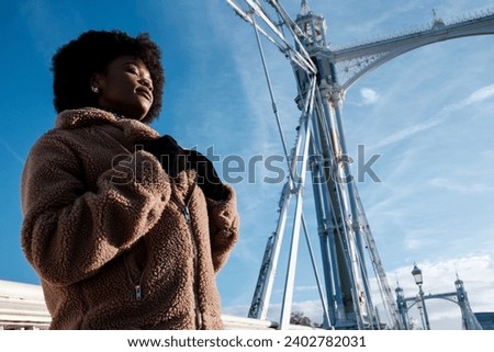 Young curly black woman enjoying winter sun under bridge structure. Her eyes are closed and she is wearing a thick coat and gloves. It is high old historical bridge in London.