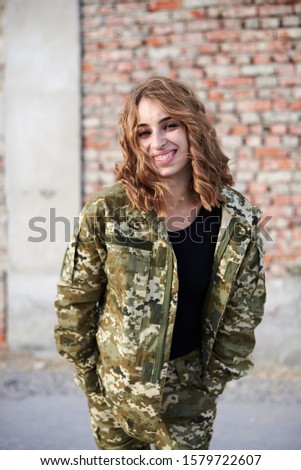 Young curly blond military woman, wearing ukrainian military uniform, posing for picture. Three-quarter portrait of female army soldier standing in front of brick wall of ruined building, smiling.