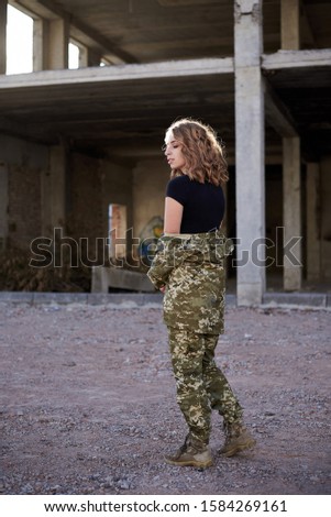 Young curly blond military woman, wearing ukrainian army military uniform and black t-shirt. Full-length portrait of female soldier walking in rim light glare in front of ruined abandoned building.
