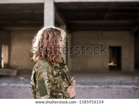Young curly blond military woman, wearing ukrainian army military uniform and black t-shirt. Three-quarter portrait of female soldier in rim light glare in front of ruined abandoned building.