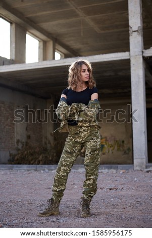 Young curly blond military woman, wearing ukrainian army military uniform and black t-shirt. Full-length portrait of female soldier standing in rim light glare in front of ruined abandoned building.