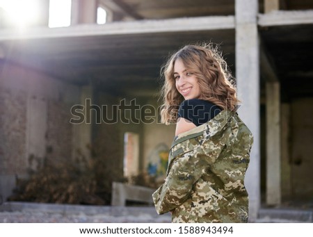 Young curly blond military woman, wearing ukrainian army military uniform and black t-shirt. Three-quarter portrait of female soldier in rim light glare in front of ruined abandoned building.
