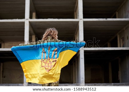 Young curly blond military woman, wearing ukrainian army uniform, holding ukrainian blue and yellow flag. Three-quarter picture from back of female soldier in front of ruined abandoned building.