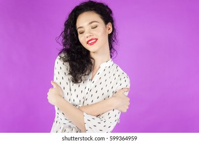 young curly brunette woman hugging herself on a pink background.