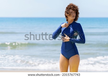 Young curly ginger haired female in blue wetsuit preparing for surfing in waving sea while looking away in sunlight 商業照片 © 