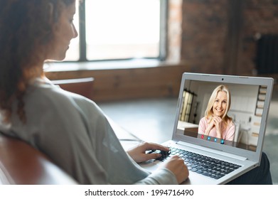 Young Curly Girl Sitting On The Couch At Home And Talking Online With Middle Aged Woman, Making Video Call To Colleague Or Mother, Talks With Psychologist Online, Takes Virtual Consultation