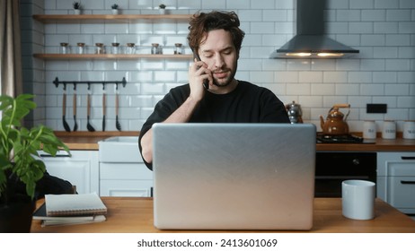 Young curly hair man sitting in the modern kitchen talking on cellphone while typing on laptop computer - Powered by Shutterstock