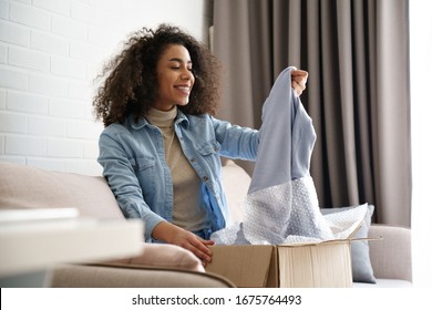 Young curly hair satisfied happy african girl woman lady shopaholic customer sit on sofa unpack parcel delivery box look at clothes take out grey pullover sweater, online shopping shipment concept.
