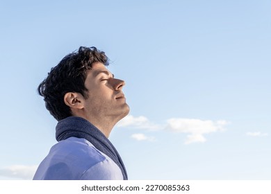 Young curly haired boy isolated on background of clear sky and with closed eyes smelling blue sky.