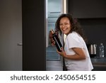 Young curly haired brunette woman with alcohol addiction, holding a bottle of wine and drinking alcohol in the morning, smiling looking at camera. Alcoholism, mental health and social problems