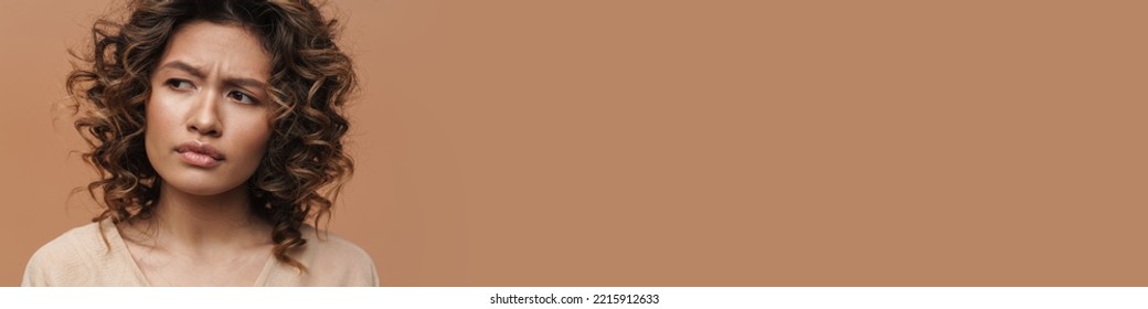 Young curly hispanic woman frowning and looking aside isolated over beige background - Shutterstock ID 2215912633